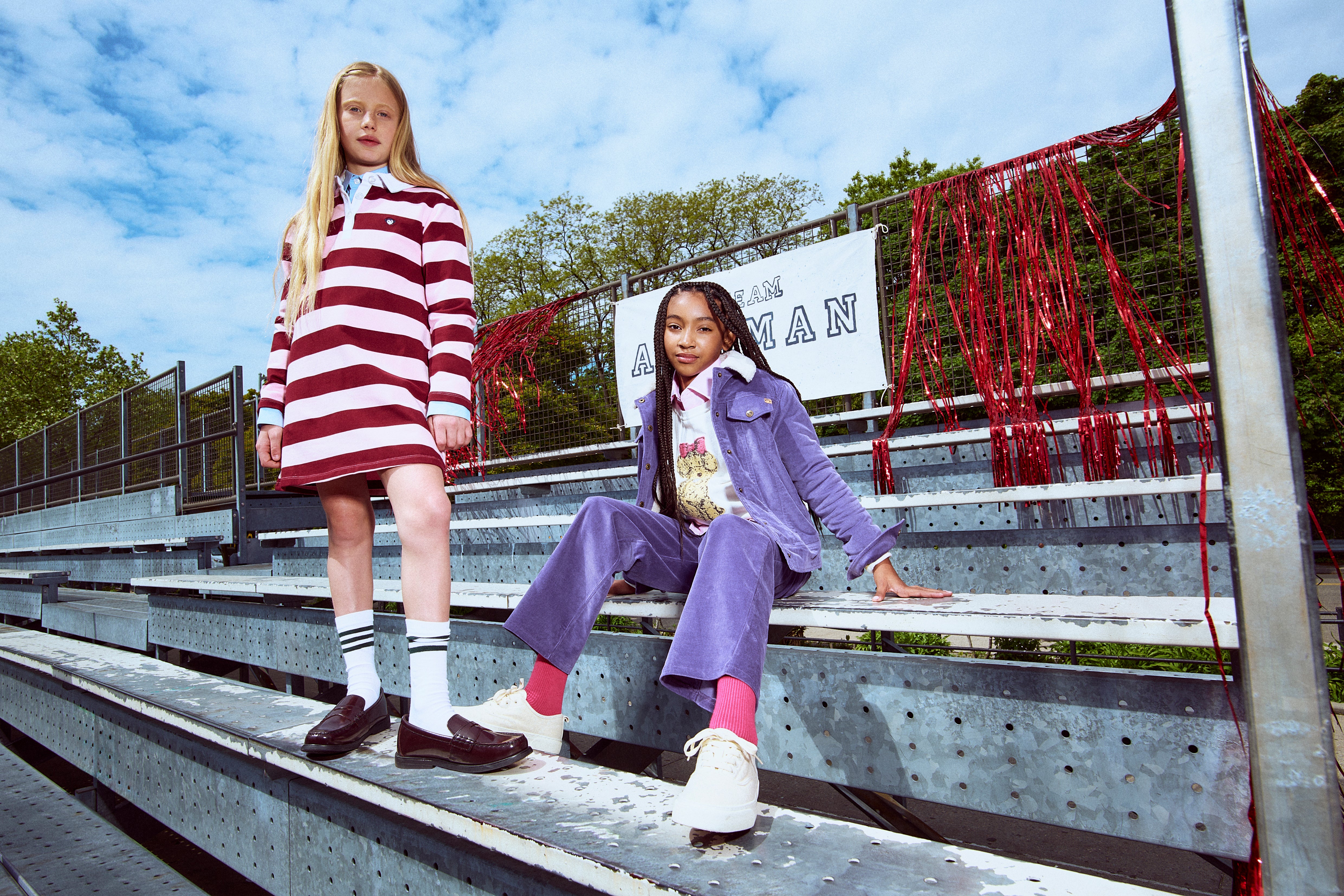 Two young models posing on outdoor metal stadium bleachers under a blue sky. The girl standing wears a red and white striped long-sleeve mini dress with white socks and brown loafers.

