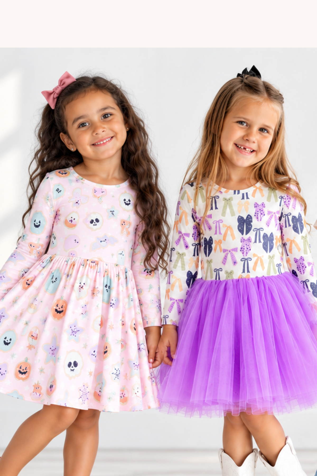 Four young girls modeling different colorful children's outfits against a bright white background with soft window shadows. The outfits include a pink long-sleeve dress with cartoon prints, a purple tulle skirt with a patterned top, a white graphic T-shirt with jeans, and a white T-shirt with a black witch graphic paired with a black skirt.