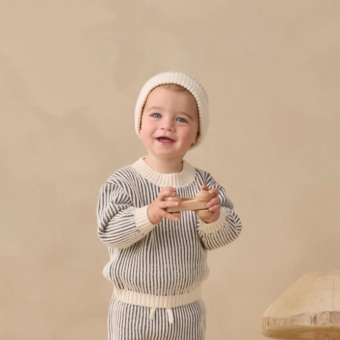A smiling toddler wearing a cream knit beanie and a cream-and-gray vertically striped knit sweater, holding a small wooden toy against a warm beige studio background.